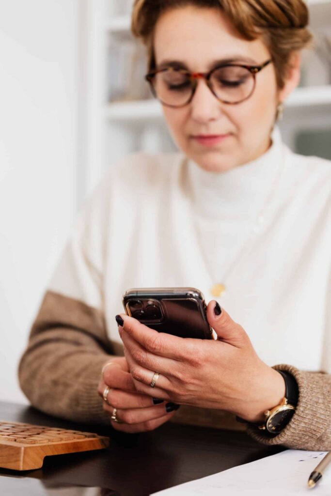 Crop content female in stylish sweater and eyeglasses surfing modern mobile phone while sitting at desk in light contemporary workspace